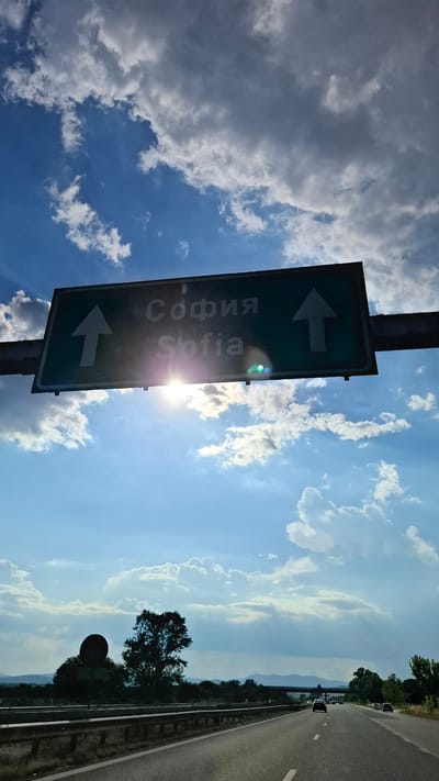 A green highway sign with white text and arrows indicates directions to Sofia. The sign is positioned above a highway. Sunlight shines brightly through the clouds, creating a lens flare on the sign and partially obscuring the text. The sky is blue with scattered white and gray clouds. Below the sign, a road stretches into the distance, with cars visible on it. Guardrails line the side of the road, and trees and other vegetation are present in the background. The scene appears to be in the vicinity of Dinkata, Bulgaria.
