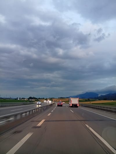 The image shows a view from a vehicle traveling on a highway, likely in or near Oberbipp, Switzerland, given the context. The sky is heavily overcast with dark, dramatic clouds. The highway has multiple lanes and is wet, indicating recent rain or damp conditions. Several vehicles are visible on the road, including cars and a white truck. The headlights of oncoming vehicles are illuminated, suggesting it is dusk or a dark day. On the sides of the highway, there are fields and distant hills or mountains. Power lines and vegetation are also visible in the background. The perspective is from the driver's point of view, looking forward.