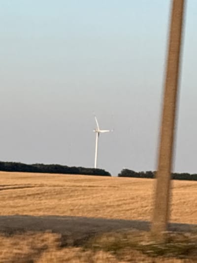 This media file captures a distant view of a wind turbine against a clear, pale blue sky. The turbine is small in the frame, with its blades appearing as a white cross-like shape. To the right of the frame, a vertical wooden structure dominates, showing a rough texture with vertical grain and some darker, possibly mossy or shadowed areas along its left edge. A thin, horizontal wooden beam stretches across the upper portion of the image, obscuring the top of the sky and partially the wind turbine. The scene appears to be outdoors, possibly in a rural or open area given the presence of the wind turbine and the texture of the wooden element. The lighting suggests it is daytime, with the sun casting shadows on the wooden structure. The overall impression is one of a wide, open landscape with industrial elements. There is no visible text in the image.