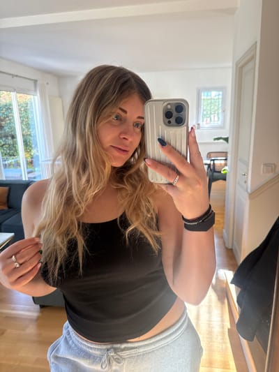 A female is depicted in a mirror selfie within a residential interior in Rueil-Malmaison, France. She possesses long, wavy blonde hair and blue eyes, and is attired in a black ribbed tank top and light grey drawstring sweatpants. Her right hand holds a silver-colored smartphone featuring a triple-lens camera array and a ribbed protective case, positioned to capture her reflection. On her right wrist, a black fitness tracker and a slender black bracelet are visible. Her right ring finger displays a silver ring with embedded stones, and her nails exhibit dark blue/black polish with light pink tips. Her left hand is raised, touching her hair, revealing two silver rings on her left ring finger (one a plain band, the other an open design) and nails with dark-colored tips.
The background, observed through the mirror, consists of white walls and light wooden flooring. To the left, a large glass door or window provides a view of dense green foliage outside, and a dark blue sofa is partially visible indoors. To the right, a white-framed window with exterior security bars shows a bright sky and distant trees. A white doorway leads into an adjacent room where a dark chair and a hanging dark garment are present. The scene is illuminated by natural light.