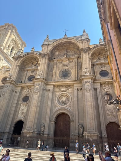 This is a low-angle, outdoor photograph of the Granada Cathedral in Granada, Spain. The sun is high in the sky, casting strong shadows on the building's ornate facade. The cathedral is constructed of light-colored stone and features numerous architectural details, including arches, columns, statues, and decorative carvings. A large wooden door is visible in the center of the facade, suggesting an entrance.
In the foreground, a group of tourists are gathered on a wide set of stone steps leading up to the cathedral. Some are looking at the building, others are taking pictures, and some are engaged in conversation. The people are dressed in casual summer clothing, and most appear to be facing the cathedral. A metal fence separates the steps from the immediate entrance area.
To the right of the cathedral, a portion of another building with a pink facade is visible, creating a strong contrast with the stone of the cathedral. The sky is clear and blue, indicating a sunny day. The overall impression is a scene of tourism and cultural exploration in a historic Spanish city.