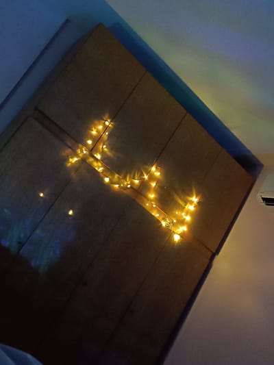 A medium shot captures a tilted view of a wardrobe or large cabinet with several vertical panels, illuminated by a string of fairy lights arranged in an irregular, non-representational pattern. The light appears warm yellow and is concentrated primarily on the right side of the object. The wardrobe is placed against the backdrop of a wall, which is light at the bottom and fades into a darker shade at the top. A corner is visible at the top-left, which shows a dark blue section of the wall. An air conditioning unit is partially visible on the right side of the image. The location is Cherkovitsa, Bulgaria.