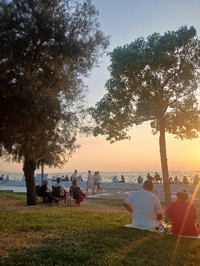 The image captures a waterfront scene in Karşıyaka, Turkey, during sunset. In the foreground, two individuals are seated on a mat on a grassy area, facing away from the viewer towards the water. To their left, beneath a large tree with dense, dark foliage, four individuals are seated in portable camping chairs. A second, slender tree with lighter green foliage stands to the right. Beyond the grassy expanse, a light-colored concrete promenade runs parallel to the water. A group of approximately five to six individuals are walking across this promenade from left to right. Numerous other people are visible seated or standing along the promenade and on a low concrete barrier or seawall closer to the water's edge. The background features a wide body of water, likely the Gulf of Izmir, with faint landmasses visible on the distant horizon. The sky displays a gradient from light blue at the top to vibrant orange and yellow near the horizon, with a significant sun flare originating from the right side of the frame, indicating the setting sun over the water.