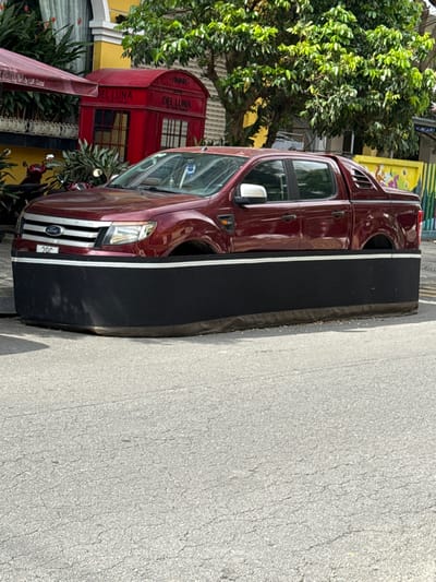 A maroon Ford Ranger four-door pickup truck, missing its wheels, is centrally positioned within a large, rectangular black planter structure with a prominent white horizontal stripe near its upper edge. The planter rests on an asphalt road surface, elevating the truck's chassis. The truck displays a "20C" license plate on its front bumper. In the background, a yellow building is visible, from which a dark red awning, labeled "DEL LUNA by LUK Global," extends. A red British-style telephone booth replica, also branded "DEL LUNA by LUK Global," stands in front of the building. Lush green trees with dense foliage are present above the building and the phone booth, casting shadows. Partially visible to the far right is a low wall with colorful abstract patterns. The scene takes place outdoors on an asphalt road with visible cracks, under bright daylight conditions in An Hải Ward, Vietnam. No individuals are visible.