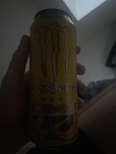 A person's hand is holding a yellow can of Monster Energy drink. The can has the Monster logo and text indicating "THE DOCTOR". The background is dark, with some light reflecting off a surface in the upper right corner. The scene appears to be indoors, possibly in a dimly lit room. There is no information in the image to suggest a location in Paczkowo, Poland.