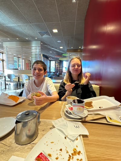 The image depicts two individuals seated at a light wooden-patterned table inside a McDonald's restaurant in Lipomo, Italy.
On the left, a young male with short dark hair, wearing a white short-sleeved t-shirt, smiles directly at the camera. He holds a clear glass containing a pale amber liquid, possibly tea, in his right hand. To his left, on a white paper tray, an orange-wrapped item is visible.
On the right, a young female with long light brown hair, wearing a dark long-sleeved top, has her eyes closed and mouth slightly open, a spoon held in her right hand near her mouth. In front of her, on a small wooden-patterned tray, are two white McDonald's cups; one contains a dark brown substance, the other is mostly empty. A white rectangular tray with a partially consumed brown pastry or cake and chocolate smears is also on this tray.
The table surface is cluttered with food remnants, including crumbs and smears, and several white McDonald's receipts and paper packets, one labeled "zucchero di canna 100% italiano" (100% Italian sugarcane). A silver metal pitcher with a lid and an empty white plate are also present on the table in the foreground.
The background shows the interior of the fast-food establishment, characterized by a red wall on the right, a white slatted partition on the left, and grey acoustic ceiling tiles with recessed lighting and air vents. Further back, windows reveal exterior greenery. Beige booth seating is visible behind the female subject.