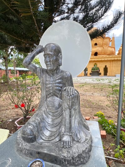 The image depicts a dark grey stone statue of an emaciated, seated figure, likely an ascetic or Buddhist monk, located outdoors within a religious complex in Đăk Mar, Vietnam. The statue, positioned centrally, shows a male figure with visible ribs, a serene expression, and a slight smile. The figure's right arm is raised, holding an elongated object (possibly a staff or brush) over its head, while the left hand rests on the raised left knee. The statue rests on a dark grey stone pedestal.
Behind the statue's head is a large, circular, white disc, possibly a halo or a decorative element. To the right of the statue, a prominent, multi-tiered golden-yellow pagoda or temple building with distinctive lotus-petal shaped structures and spires is visible in the midground. Archways at the base of the temple reveal dark silhouetted figures or architectural details within.
Overhead and to the left, dark green foliage from a large evergreen tree partially shades the scene. The foreground and midground consist of bare earth with sparse green vegetation, including small bushes with red flowers (like a rose) to the left and yellow flowers to the right. Red bricks are scattered on the ground near the base of the statue on the right. In the immediate foreground, a small blue and white patterned bowl containing a dark substance, possibly ash, rests on the statue's base. The background features a partly cloudy sky. To the far left, a simple building with a red roof is partially visible amidst trees.