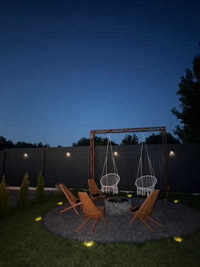 The image depicts an outdoor seating area at dusk in Sharovechka, Ukraine. The scene features a circular gravel pit with a fire ring at its center, surrounded by four wooden Adirondack chairs. Two white net swings hang from a wooden A-frame structure behind the fire pit. A dark metal fence runs along the background, with small lights illuminating it at intervals. The lawn in the foreground is lit by several small, yellow ground lights. Trees and foliage are visible behind the fence, and the sky is a deep twilight blue with a few visible stars. No people are present in the image.