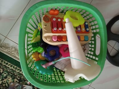 A green plastic laundry basket filled with children's toys sits on a tiled floor. The toys include a toy xylophone/keyboard with multicolored keys and an orange button, a white plastic toy with a conical shape and a blue tubing, and multiple colorful toy rings. Part of a patterned rug is visible to the left of the basket, and part of a black wheel-like object is to the right. The image appears to have been taken from an elevated position, looking down into the basket. Location: Palohigeuh, Indonesia.