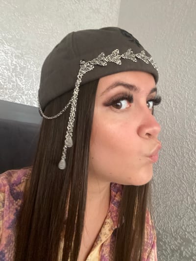 A young woman with dark hair is looking to her right and puckering her lips. She is wearing a dark-colored beret adorned with silver metal scrollwork and hanging chains with teardrop-shaped charms. Her makeup includes long, false eyelashes and winged eyeliner. She is wearing a colorful shirt with a pattern of purple and yellow shapes. The background appears to be a textured gray wall. There is no indication of the location being Chalda, Russia.