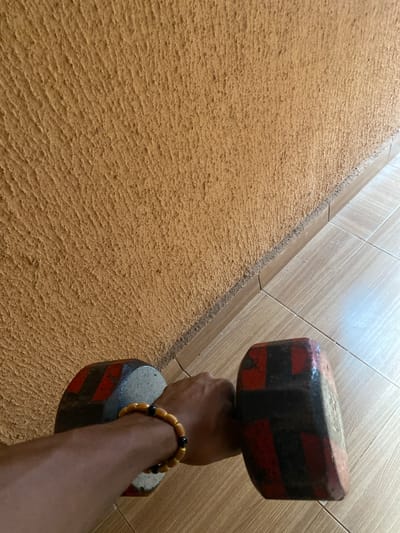 The image shows a close-up, over-the-shoulder perspective of a person's arm and hand holding a dumbbell. The hand is adorned with a beaded bracelet featuring yellow and black beads. The dumbbell is heavy and appears to be made of metal, with red and black markings on its hexagonal ends. The background consists of a textured, orange-brown wall and a section of light-colored tiled flooring. The setting is indoors, possibly in a home or gym, and is not specifically identifiable as being in Amaokpala, Nigeria, based on the visual information provided.