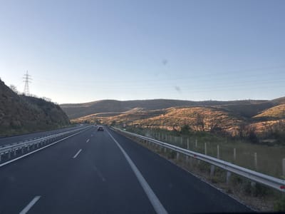 An outward-facing drone shot captures a highway with three lanes, two heading in the same direction, as it cuts through a valley in Avra, Greece. The road is dark asphalt, with a white line separating the lanes and a solid white line on the far left. A silver guardrail runs along the right side of the road. The landscape on both sides of the highway is a dry, hilly terrain covered with sparse, brown vegetation. In the distance, rolling hills are illuminated by the golden light of the setting sun, creating a warm glow. Power lines and a tall electrical tower are visible on the left side of the frame. There are no people visible, but two cars are driving on the highway. A circular speed limit sign with the number 90 is visible on the right side of the road.