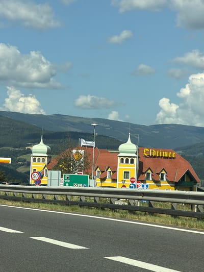 The image captures a roadside view of a building with a prominent "Oldtimer" sign in Maierhöfen, Austria. The building features a distinctive yellow facade with red-tiled roofs and green-domed towers. Various road signs are visible, including a speed limit sign of 50 and a STOP sign, indicating a nearby intersection or junction. A green sign with symbols for a gas station and parking is also present. The scene is set against a backdrop of rolling green hills and a partly cloudy blue sky. The foreground shows a section of asphalt road with white lane markings and a metal guardrail. There are no discernible people in the immediate vicinity of the building or on the road. The overall impression is that of a service area or rest stop along a highway.