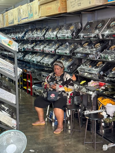 In Ranong, Thailand, a woman sits on a black stool in what appears to be a retail store or warehouse. She is wearing a black headband, a patterned long-sleeved shirt with black, white, and red colors, and black shorts. She holds a smartphone in her hands and has a small denim purse with a pink flower emblem slung over her shoulder. The store is filled with rows of new gas stoves, each packaged in clear plastic. The shelves are stocked high with these appliances. In the foreground, a white fan is visible on the floor. The environment suggests a commercial setting with a focus on kitchen appliances.