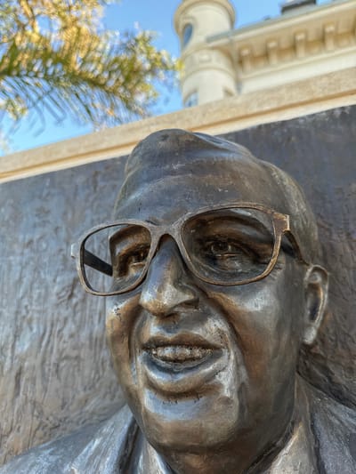 The image shows a close-up of a bronze bust of Jorge Luis Borges, identified by his distinctive glasses. The bust is mounted on a wall, and the glasses reflect the blurred image of trees and a building with architectural detail. The bust is located in Tigre, Argentina.