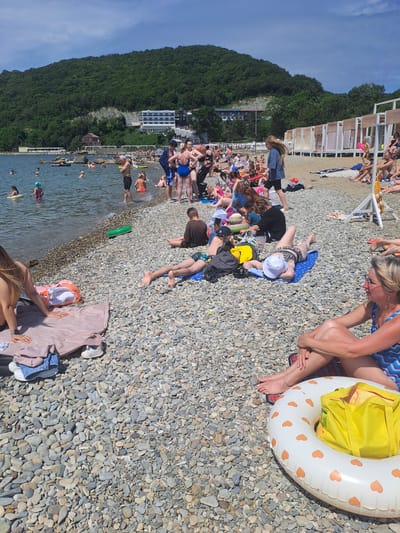 The image depicts a crowded pebble beach in Dzhubga, Russia, under a clear blue sky with scattered clouds. The water is calm and clear, with several people wading and swimming. On the beach, numerous individuals are sunbathing, sitting, or walking among the pebbles. Beach tents or cabanas are set up in the background, providing shade. The overall scene is one of a busy summer day at a seaside resort.