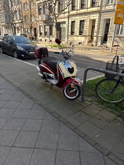 An urban street scene in Aachen, Germany, depicted on a clear day with direct sunlight. In the immediate foreground, a two-tone red and cream vintage-style motor scooter, featuring a windshield, dark brown seat, and rear storage box, is parked on a grey tiled sidewalk. Its front wheel, characterized by a whitewall tire and red hub, is secured by a black chain lock to a grey metal bicycle rack. A dark blue bicycle with a front basket is partially visible, also parked and secured to the same rack. Adjacent to the sidewalk, a dark grey Peugeot 208 hatchback with a Belgian license plate "1-DNO-1" is parked parallel to the curb on an asphalt street. In the background, multi-story buildings with light-colored facades and numerous windows line the street, interspersed with bare deciduous trees. A yellow directional street sign on the right indicates "Burtscheid," "Vaals," and "Uniklinik." No individuals are present in the observable scene.
