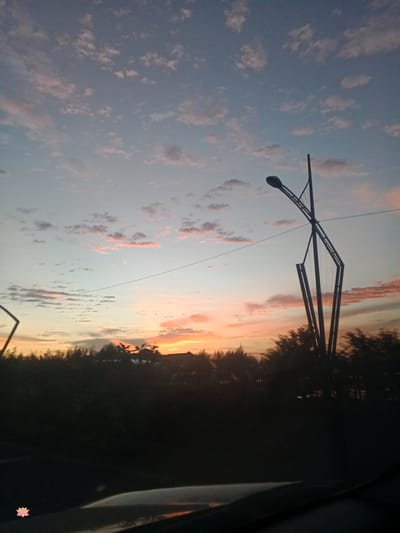 The image depicts a sunset scene with a street lamp in the foreground, silhouetted against a colorful sky. The sky is filled with scattered clouds tinged with pink and orange hues, indicating the setting sun. A thin power line stretches horizontally across the frame. In the background, a dark silhouette of trees and some structures is visible. The lower part of the image shows a dark surface, likely part of a vehicle, and a small pink lotus flower graphic is present on the left side. The scene is set in Cotkareung, Indonesia.