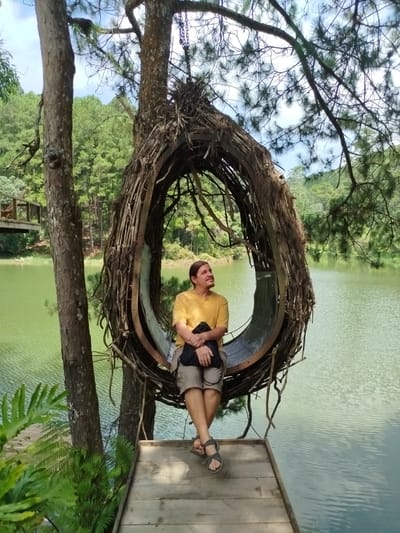 The image captures a scenic view of Cam Ly waterfall in Da Lat, Vietnam, with a unique bird's nest-shaped swing as the focal point. A person, wearing a yellow t-shirt and khaki shorts, is seated on the swing, which is suspended from a tree by ropes. The swing is made of natural materials, resembling a large nest woven from branches and straw. The person is looking upwards, seemingly enjoying the peaceful ambiance.

The background features a lush green forest, typical of Da Lat's mountainous terrain, with a calm, greenish lake in the foreground. The water reflects the surrounding trees and the sky, creating a serene and tranquil atmosphere. A wooden platform is visible beneath the swing, suggesting it's part of a tourist attraction. The overall scene depicts a blend of natural beauty and creative design, offering a picturesque spot for relaxation and photography.