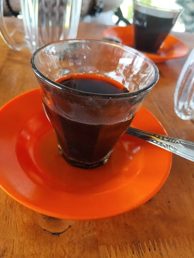 A close-up, eye-level shot shows a clear glass tumbler filled with dark liquid, likely coffee, sitting on a bright orange saucer. The tumbler has a ridged texture and is positioned slightly off-center to the left of the frame. A silver spoon rests on the saucer to the right of the tumbler, its handle extending out of the frame. The orange saucer is on a wooden table with visible grain and some dark imperfections. In the background, out of focus, is another orange saucer with a similar tumbler of dark liquid, and a clear glass mug with a handle. The setting appears to be outdoors, with blurred green foliage and bright light suggesting a sunny day. This scene is likely located in Meunasahmee, Indonesia, given the context.