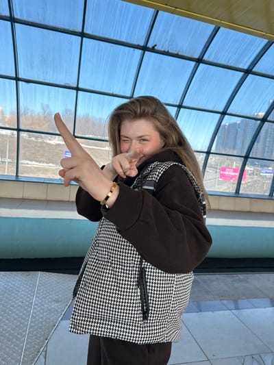 A female subject is depicted posing within a glass-enclosed pedestrian bridge or covered walkway in Nikol'skii, Russia. The subject, with light brown hair, is positioned slightly right of center, facing the camera. She is winking with her right eye and smiling. Her left arm is raised, with the index finger extended upwards and to the left, while her right hand is also raised, with the index finger extended forward. She wears a dark brown hooded garment under a sleeveless vest featuring a black and white houndstooth pattern. A bracelet with alternating gold-colored and black rectangular segments is visible on her left wrist.
The environment features a curved, glass-paneled roof structure with dark frames, through which a clear blue sky is visible. Beyond the structure, bare trees and several multi-story buildings are discernible in the distance. Below, a road or thoroughfare is visible, including a prominent pink billboard displaying white Cyrillic text that reads "РУКИ ВВЕРХ" (HANDS UP). The floor of the walkway consists of grey tiles and a section of metallic diamond plate flooring. A horizontal light blue ledge or barrier is present along the lower section of the glass panels.