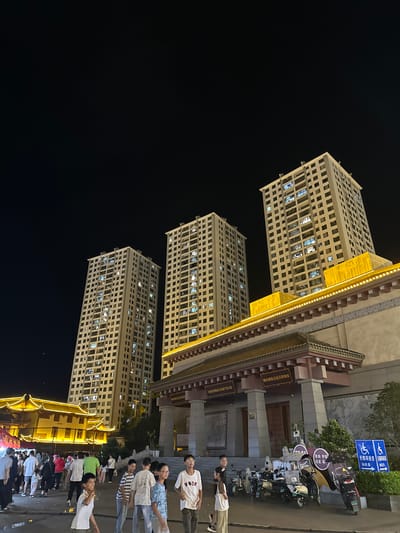 In Eshan Yi Autonomous County, China, at night, the scene depicts a lively outdoor area in front of buildings. Three tall, light-colored residential buildings with numerous illuminated windows stand against a dark sky, suggesting they are apartment complexes. In the foreground, a traditional Chinese-style building with a dark, ornate roof and bright yellow decorative lighting runs across its facade. People are gathered in the open space in front of this building. Some individuals are walking, while others appear to be standing and conversing. Several scooters and motorcycles are parked on the right side of the image, near a pathway. Blue signs with white symbols, indicating accessibility for people with disabilities, are also visible on the right. The overall atmosphere appears to be that of a public gathering or market at dusk or nighttime.
