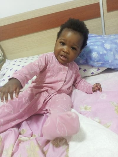 A baby, wearing pink pajamas with white bear prints, sits on a bed. The baby has dark hair and skin, with a slightly open mouth and focused gaze. A light pink blanket with a floral pattern covers the bed, and a white and blue pillow is behind the baby. The headboard is partially visible in the background.