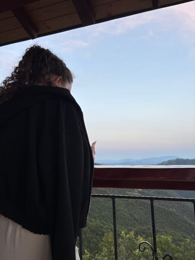 A person with curly dark hair is standing on a balcony, facing away from the camera and looking out at a mountainous landscape. The person is wearing a black hooded jacket and a light-colored garment. The balcony has a wooden railing and a metal railing below it. The sky is light blue with some wispy clouds, and the sun appears to be setting or rising, casting a soft light on the mountains in the distance. The location is described as being in the city of Pechinska, Bulgaria, and the image captures a serene natural scene.