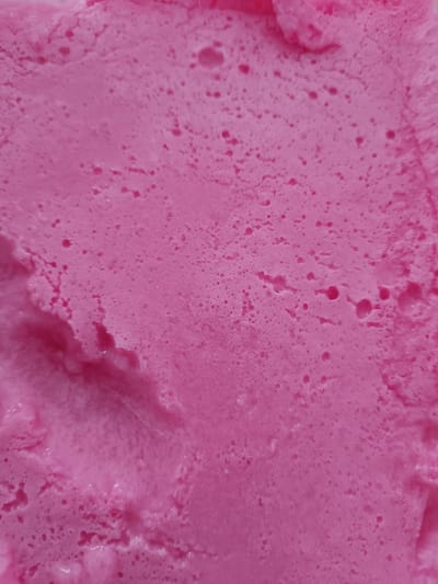 The image is a close-up, top-down view of a pink, frothy substance, likely ice cream or a similar dessert. The texture is uneven, with many small air bubbles and some larger indentations and ridges. The color is a vibrant pink, with slight variations in shade. There are no discernible objects, people, or actions within the frame. The image does not provide any information about its location in Vitoria-Gasteiz, Spain.