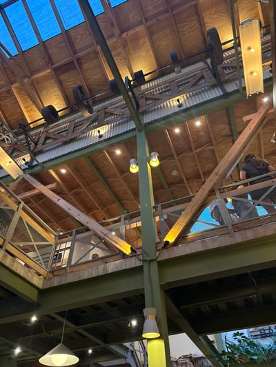 The image depicts a low-angle view of a building's interior, possibly a commercial space or restaurant, in San Cristóbal, Mexico. The architectural structure features an exposed wooden beam ceiling with sections of corrugated metal roofing. Industrial-style lighting fixtures are present, including exposed bulb lamps and hanging cone lights. A decorative arrangement incorporating repurposed industrial items, such as metal wheels or gears, is suspended from the ceiling. The building structure also includes green-painted metal beams and a multi-level design with metal railings and grating, suggesting a mezzanine or platform area. An individual or individuals can be seen on one of the upper levels. There are also potted plants and other decorative elements in the space.