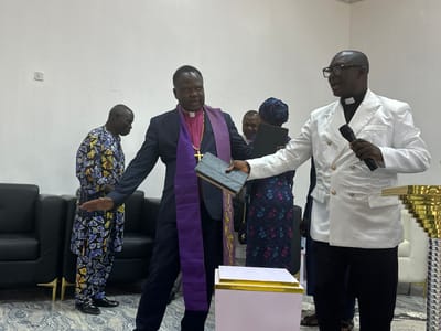 This image captures an indoor gathering within a brightly lit room in Goni Gara, Nigeria, featuring five discernible individuals and various objects. The room features plain white walls, with two white electrical outlets visible on the left, and a patterned grey and white tiled floor.
From left to right:
A dark-skinned man with short grey hair is standing, oriented towards the center. He wears a full-length, two-piece traditional garment with a repeating blue, yellow, and black geometric pattern, paired with black patent leather dress shoes. His left arm is extended forward at waist height, palm flat and facing downwards.
Adjacent to him, a dark-skinned clergyman with short dark hair stands facing slightly right. He wears a dark suit jacket over a pink clerical shirt with a white clerical collar, and a purple stole draped around his neck. A prominent gold chain with a crucifix pendant hangs from his neck. He holds a dark-colored book in his left hand and extends his right arm forward.
In the immediate foreground, a light pink rectangular pedestal with gold trim on its edges is positioned centrally.
To the right of the central clergyman, another dark-skinned clergyman with a bald head and black-framed glasses stands facing left. He is attired in a white double-breasted suit jacket with four gold buttons on the visible side, worn over a black clerical shirt with a white clerical collar. He holds a black microphone in his right hand, appearing to be addressing the individuals to his left. His left hand is partially visible, extended towards the central clergyman.
In the background, between the two clergymen, a dark-skinned man in a white shirt is partially visible. To his right, a dark-skinned woman wearing a blue floral-patterned dress and a matching headwrap holds a dark tablet or book.
Furnishings include two black leather armchairs on the far left and a partially visible white upholstered armchair on the far right. A tall, golden, multi-tiered decorative stand is also visible on the far right.