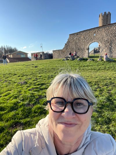 The image presents a close-up selfie of an adult female in the foreground, positioned outdoors under a clear blue sky in Chambles, France. The woman has short, light blonde hair, styled with bangs and visible flyaways at the crown. She wears large, dark tortoiseshell-patterned eyeglasses and a light beige quilted jacket, displaying a closed-mouth smile.
Behind the woman, a vibrant green grassy slope extends into the midground. To the left, a light-colored building with a dark roof and an unreadable sign is partially visible, with coniferous trees in the background. A solitary individual stands on the grassy slope near this building. A black, curved streetlamp stands to the left of a prominent stone wall.
The central background features a substantial stone wall, identifiable as part of the Château de Chambles, which includes an arched gateway. A tall, cylindrical stone tower with crenellated battlements rises behind the wall on the far right. Multiple individuals are present in the background: two people are seated on rocks near the wall to the left of the arch, another person wearing a pink/red hat is seated on the right, and several other figures are standing or moving near the wall and through the archway. The scene is brightly illuminated by direct sunlight, casting distinct shadows on the grass.