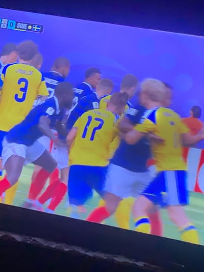 The image displays a soccer match, as indicated by the score and team names visible in the upper left corner: "0 SWE 0". Players in yellow and blue jerseys are engaged in gameplay. One player in a yellow jersey with the number "3" and the name "LINDELOF" on the back is partially visible. Another player in a yellow jersey with the number "17" is also present. Players in blue jerseys appear to be on the opposing team. The background is a solid purple, suggesting a digital or projected environment, not a real-world stadium in Zastavna, Ukraine. The scene captures a moment of physical interaction between players, typical of a soccer game.