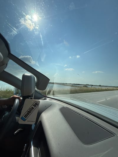 The image is a first-person perspective shot from inside a car, looking out through the windshield. The photograph is rotated approximately 90 degrees counter-clockwise.
**Scene and Setting:** The perspective is from the driver's or passenger's seat, capturing both the car's interior and the exterior landscape through the front window. The overall setting is a sunny day on the road, likely in Skutare, Bulgaria, as provided by the context.
**Subjects - Objects:** In the foreground (bottom right of the rotated image), a hand is visible gripping the steering wheel. A grey air freshener, rectangular in shape, hangs from the rearview mirror. Parts of the dashboard, including a speaker grille, are visible. The rearview mirror assembly is prominent. Through the windshield, a wide, bright blue sky with scattered white clouds dominates the upper left. The sun, intense and low in this part of the frame, causes significant lens flare and reflections on the windshield, indicating a bright day and possibly a dirty or smudged window. A long, straight road with a white lane marking runs parallel to what appears to be a body of water (possibly a river or canal) lined with vegetation.
**Activity or Event:** The visible hand on the steering wheel indicates that the car is being driven or is about to be driven, suggesting a journey or travel.
**Notable Details:** The time of day is bright daylight, likely midday or early afternoon, given the high intensity of the sun. The weather is clear and sunny with light cloud cover. The landscape features a road alongside a water body.
**Visible Text:** On the air freshener hanging from the rearview mirror, the text "mobile.bg" is clearly visible in blue lettering on a white background. Below it, in smaller script, "www.mobile.bg" can also be read. The text is rotated along with the image, appearing upside down relative to its normal reading orientation.