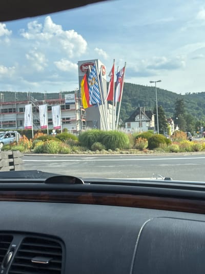 This is a photograph taken from inside a vehicle, looking out at a street scene in Bürgstadt, Germany. The image is oriented vertically. On the left, there are flags flying. One flag is a blue and white checkered pattern. Another flag is the German flag, with black, red, and yellow horizontal stripes. Behind the flags, a building is under construction, with scaffolding and white banners. To the right of the building and flags, there is a road with a single lane. Lush green trees and bushes line the side of the road. In the background, there are more trees and some buildings on a hillside. The sky is blue with white, fluffy clouds, suggesting it is daytime and the weather is fair. The interior of the car is visible in the foreground on the right, showing dark grey plastic and wood trim, and an air vent. There is a reflection of the outside scene on the car's window. Some text is visible on the white banners on the building, but it is difficult to read clearly. One banner appears to have a logo at the top.