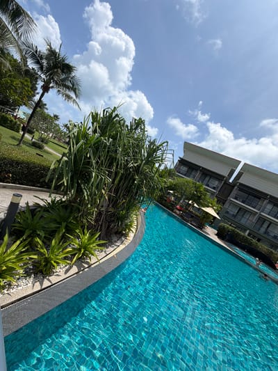 A wide shot captures a large, vibrant turquoise swimming pool with tiled bottom, reflecting the bright blue sky and scattered white clouds. The pool is situated in a resort setting, with lush green landscaping and palm trees flanking the left side. In the background, a modern resort building with multiple floors and balconies is visible. Several people are in the pool, some swimming, others relaxing. The image is taken from a slightly elevated angle, with the foreground dominated by the pool and the surrounding vegetation. The overall atmosphere is one of a sunny, tropical vacation. This image is located in Bang Muang, Thailand.
