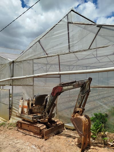 An old, rusty excavator sits in front of a large greenhouse structure. The greenhouse has a metal frame covered in translucent plastic sheeting, which appears weathered and somewhat dirty. A white pipe runs horizontally across the front of the greenhouse, with a downspout visible on the left. The ground in front of the excavator is dirt and gravel, with some sparse green vegetation to the right. The excavator is green and red, with visible rust and wear. The brand name "MITSUBISHI" is visible in white text on the arm of the excavator. The sky above is partly cloudy with patches of blue sky visible, suggesting it is daytime. The overall scene depicts an outdoor agricultural or construction setting, possibly in a rural or semi-rural area, given the greenhouse and excavator. The presence of the aged equipment and the greenhouse hints at agricultural activities, possibly in a developing or cost-conscious environment. The lighting suggests it is mid-day, with natural light illuminating the scene.