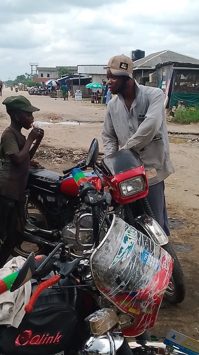 In Warri, Nigeria, a young boy in a green cap and brown t-shirt stands near a red and black motorcycle, eating something from a packet. He is facing the motorcycle, with his back slightly towards the viewer. To his right, a man wearing a grey shirt and a tan baseball cap stands leaning against the motorcycle, looking towards the boy. He has a beard and appears to be speaking or interacting with the boy. The motorcycle is the central focus, with its red body, black seat, and chrome accents. It has a clear windshield with stickers on it. The background shows a dusty, unpaved road lined with buildings and stalls, typical of a market or roadside area in Warri. There are other motorcycles and some people visible in the distance. The sky is overcast, suggesting a cloudy day.