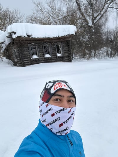 A selfie shows a person with a "Torero" printed neck gaiter and a blue jacket against a snowy backdrop. To the left, a weathered, snow-covered wooden building with broken windows stands amidst barren trees. The ground is completely blanketed in snow, creating a stark, wintery scene.