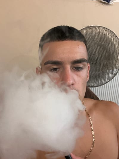 A man with a buzz cut and dark eyebrows exhales a cloud of white vapor, partially obscuring his face. He is wearing a gold chain necklace. Behind him, a circular fan with a mesh grill and striped vertical blinds are visible. The background is a plain, light-colored wall. The image appears to be taken indoors, likely in a residential setting. There is no visible indication of the specific location within the city of Каменар, Bulgaria.