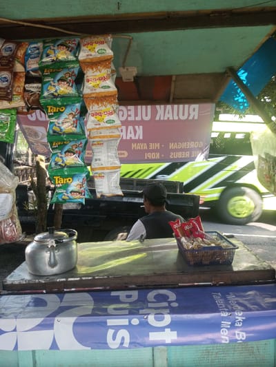 Here's a concise description of the image:

The photo appears to be taken from a food stall or mobile vendor location. Hanging from the stall roof are multiple bags of snacks. The snacks consist of bags of seaweed flavored Taro chips, and Rin-bee brand snacks. There is also a tea kettle and some snacks on the counter, and in the middle of the photo there is a man wearing a black hat sitting behind the counter facing away. A vehicle is blurred in motion on the road outside the stall. The text on the banner behind the man is in a foreign language. The bottom of the counter also has promotional material. There is no indication or evidence the image was taken in Nhot Chang, Laos.