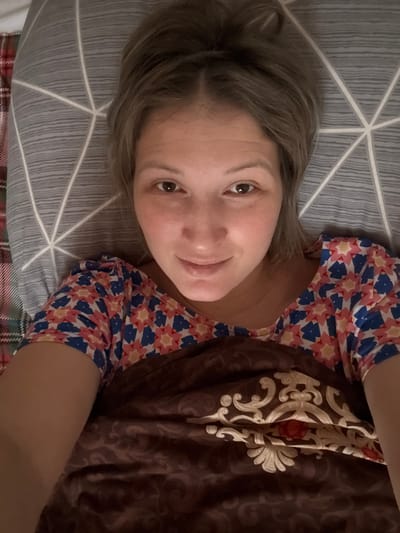 A fair-skinned woman lies on a patterned pillow, angled towards the camera. She has light brown hair and a gentle smile. Her colorful floral-patterned shirt peeks from under a brown, intricately patterned blanket or comforter. The background consists of a gray pillow with geometric white lines, and a hint of a red and green plaid fabric is visible beneath the pillow.