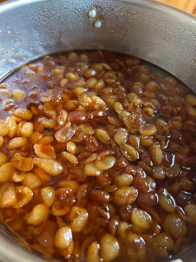 Inside a metallic pot, likely stainless steel, numerous rehydrated dried fruits are submerged in a translucent amber-brown liquid. The fruits vary in color from golden yellow to reddish-brown, appearing plump and softened, consistent with rehydrated raisins or similar dried produce. The liquid surface shows reflections of ambient light. The inner rim of the pot above the liquid line is covered with fine condensation droplets, indicating the presence of steam or a temperature differential. Two silver-colored rivets are visible on the upper interior wall of the pot. The pot is situated on an indistinct, light-colored surface, possibly a wooden countertop, in the city of Konstantinovo, Bulgaria. No individuals or specific actions are depicted.