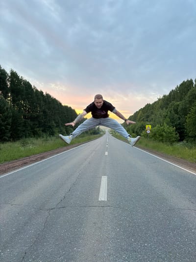 A man is captured mid-air, performing a wide split jump on an empty asphalt road. He is wearing a black t-shirt, light blue jeans, and white sneakers with yellow accents. The road stretches into the distance, flanked by dense green forests on either side. The sky above is a mix of grey clouds and a soft orange hue from the setting sun. A yellow diamond-shaped road sign with a black border and a black symbol of a pedestrian with a child is visible on the right side of the road, with a circular sign below it indicating "70". The scene appears to be in a rural or semi-rural area, possibly on the outskirts of Новая Казмаска, Russia, given the presence of natural landscape and a paved road.
