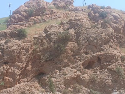 The image depicts a rugged, rocky mountainside with sparse vegetation. The terrain is uneven and appears to be composed of weathered rock formations. Small shrubs and dry grasses are scattered across the slopes. In the background, a clear blue sky is visible, and a telecommunications tower can be seen on the left side of the frame. The scene appears to be an outdoor, natural environment, possibly in the mountainous region of Farab, Tajikistan. There are no people or overt signs of human activity other than the distant tower.