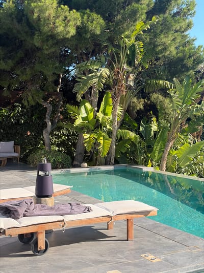 In the city of Vouliagmeni Municipal Unit, Greece, a serene outdoor scene unfolds by a shimmering turquoise swimming pool. The pool's edge is lined with grey stone tiles, reflecting the lush greenery that surrounds it. Two wooden lounge chairs, each topped with a cream-colored cushion, are positioned on the tiled patio. One lounge chair features a folded grey towel and a dark cylindrical speaker resting on a wooden block. In the background, tall pine trees and large-leafed tropical plants create a vibrant, verdant backdrop under a clear blue sky. A single armchair with a light-colored cushion is visible on the far left, adding to the tranquil atmosphere of a relaxing vacation spot.