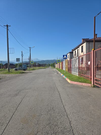 The image depicts a street scene in the city of Ir, Russia, on a clear, sunny day. A paved road runs through the center of the frame, leading towards a distant mountain range. On the right side of the road, a colorful metal fence borders a building with a light-colored facade and a brown roof. The fence is decorated with alternating sections of red, yellow, white, and decorative metalwork. Several pedestrian crossing signs are visible along the road. On the left side of the road, there are utility poles with overhead wires, a car parked on the grassy shoulder, and some houses in the background. The environment appears to be a semi-urban or rural area, with elements of both residential buildings and natural landscape.