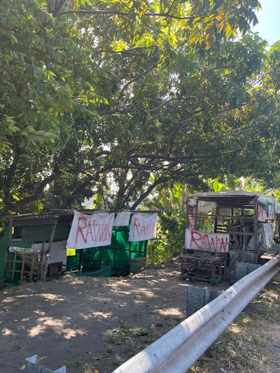 The image depicts an outdoor scene with two makeshift structures set up along a road, possibly a roadside stall or shelter. The structures appear to be constructed from scrap materials like wood, metal, and tarpaulin, with one of them covered by a green tarp on the left and the other by a grey tarp on the right. White banners with red lettering are hung on both structures. The banner on the left appears to read "RAWAN" (meaning "danger" in Indonesian). The banner on the right also seems to display "RAWAN".

The area around the structures is unpaved, with dirt and scattered leaves on the ground. A metal guardrail runs along the edge of the road in the foreground, separating it from the area where the stalls are located. Lush green foliage from large trees overhangs the scene, creating dappled shade on the ground. In the background, beyond the structures, there are more trees and vegetation, suggesting a semi-rural or vegetated roadside environment. The sky is visible through the leaves, indicating it is daytime, likely sunny given the bright lighting and shadows.

The image does not contain any people or visible interactions. The overall impression is one of a simple, possibly temporary, setup in a vegetated roadside area. The presence of the "RAWAN" banners suggests a warning or concern about something in the immediate vicinity. Without further context, the specific location in Pamotan, Indonesia, cannot be definitively confirmed from the visual information alone, but the presence of Indonesian language on the banners makes this location plausible.