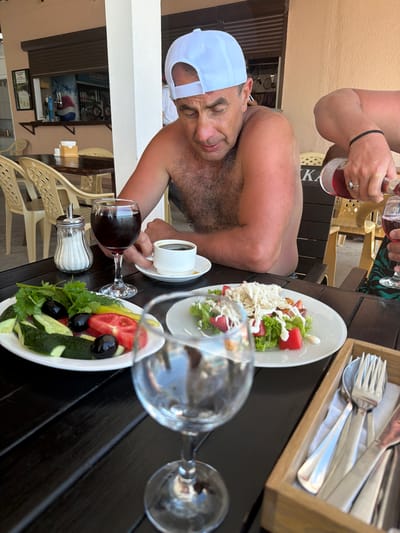 The image depicts a man wearing a white baseball cap, shirtless and looking down at his food on a table outside a restaurant in Tsandrypsh, Abkhazia. The table is set with plates of salad, a glass of red wine, a sugar dispenser, and a cup of coffee. A person is pouring wine from a bottle into a glass. The background shows a bar area with a large cooler and other restaurant seating. The overall scene suggests a meal being enjoyed outdoors.