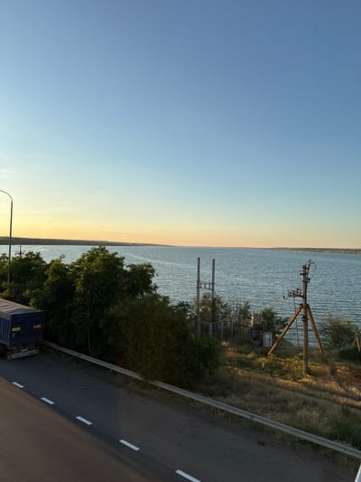 The image depicts a wide body of water, likely a river or reservoir, under a clear blue sky with a faint yellow glow near the horizon, suggesting either sunrise or sunset. The water has a slightly rippled surface. On the left side of the frame, a dark blue truck with a white tarp is parked on an asphalt road that runs parallel to the water. The road has white dashed lane markings. A metal guardrail separates the road from a grassy embankment. Dense green trees and bushes line the embankment, obscuring some of the view of the water. On the right side of the embankment, there are several utility poles with electrical equipment and some low-lying structures. The background shows the far bank of the water body, which appears to be a natural landscape with some vegetation. The overall scene is peaceful and natural, with a hint of industrial infrastructure. The location is identified as Yelyzavetivka, Ukraine.